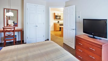 A hotel room with a bed, a television, a dresser, a chair, a desk, and a mirror. There is a hallway leading to a kitchen area.