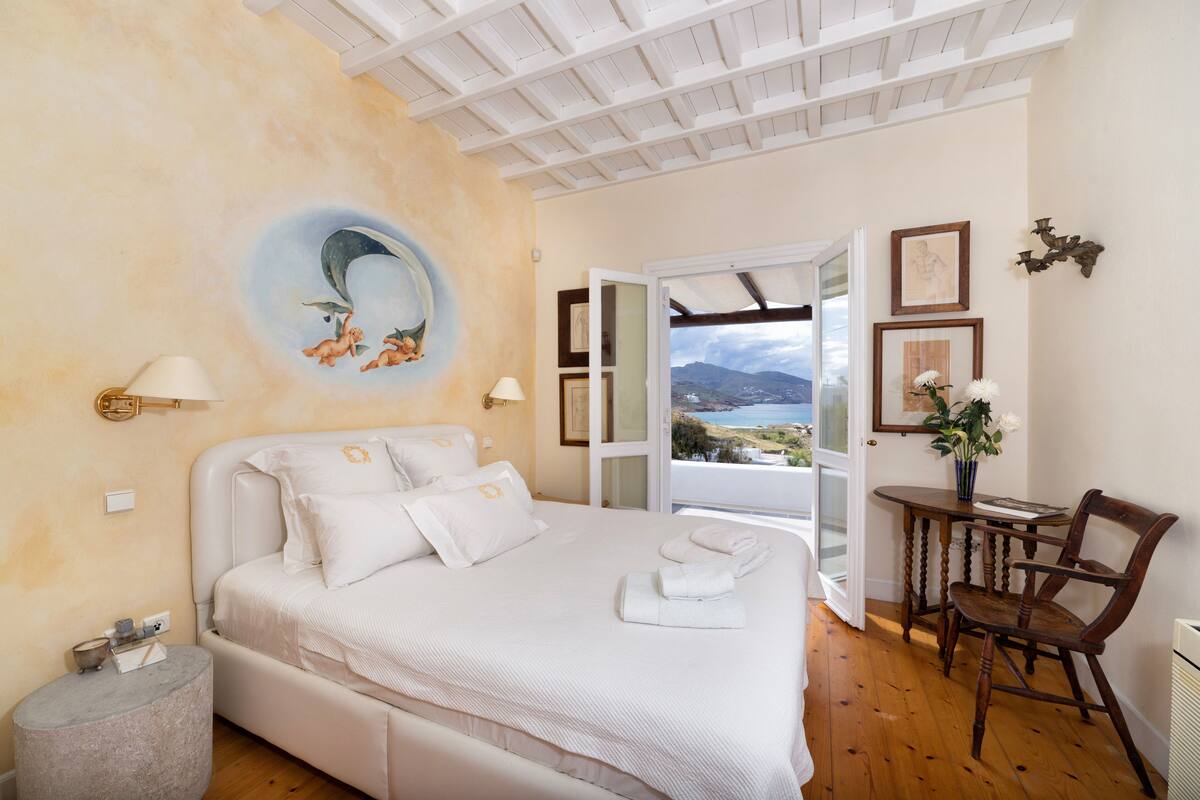 A bedroom with a bed, a round wall decoration, a table with a chair, and a view through an open door.
