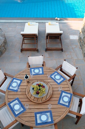 A round wooden table with blue placemats and white cushions, surrounded by chairs, set on a patio with a pool in the background. - Villa Galaxy (Mykonos)