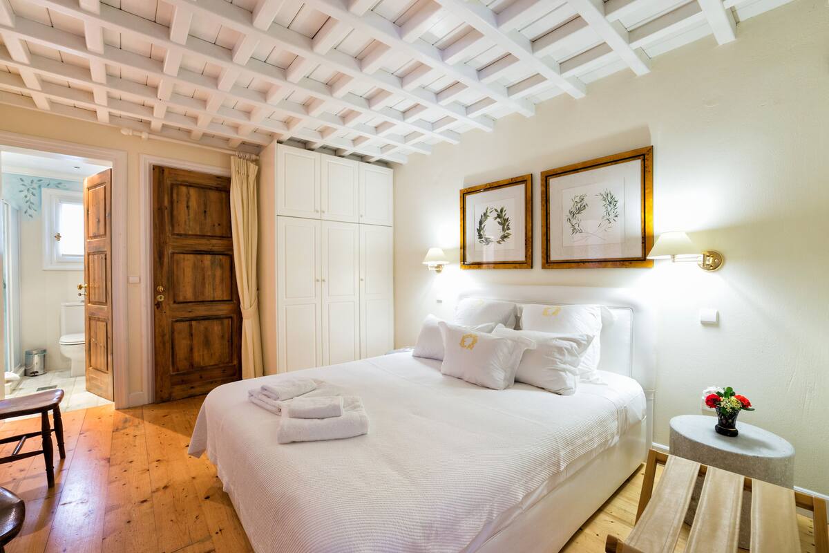 A bedroom with a bed, white curtains, a wooden door, a small round table with a vase, and framed pictures on the wall.