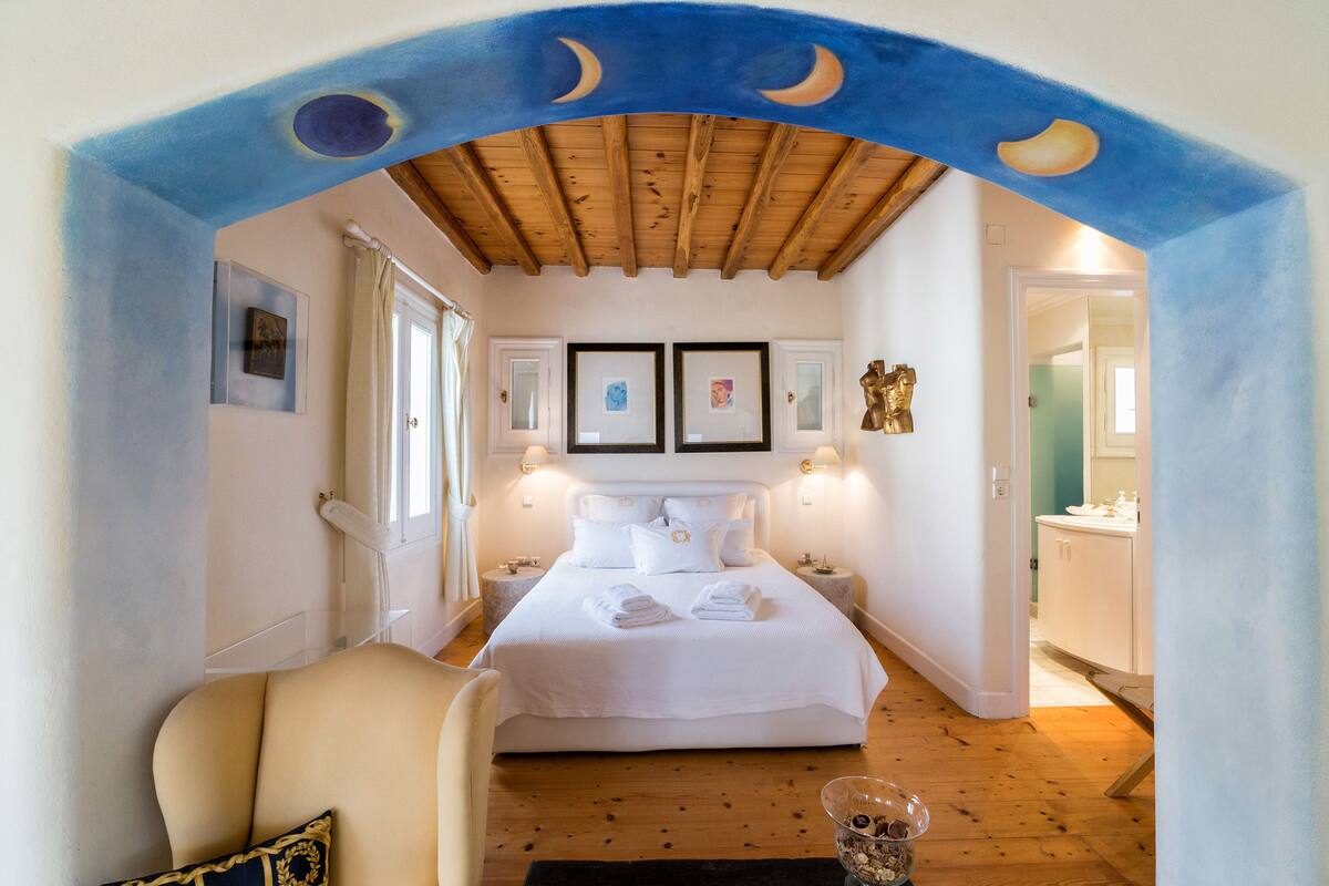 A bedroom with a wooden ceiling, a bed with white linens, two framed pictures on the wall, and a nightstand with a lamp.