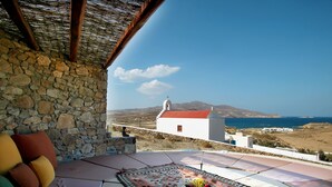A stone wall, a white building with a red roof, and a view of the sea and hills. - Villa Galaxy (Mykonos)