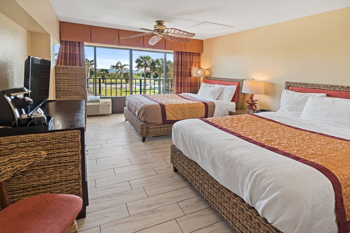 A hotel room with two beds, a desk, a chair, a television, and a balcony with a view of palm trees and the ocean.