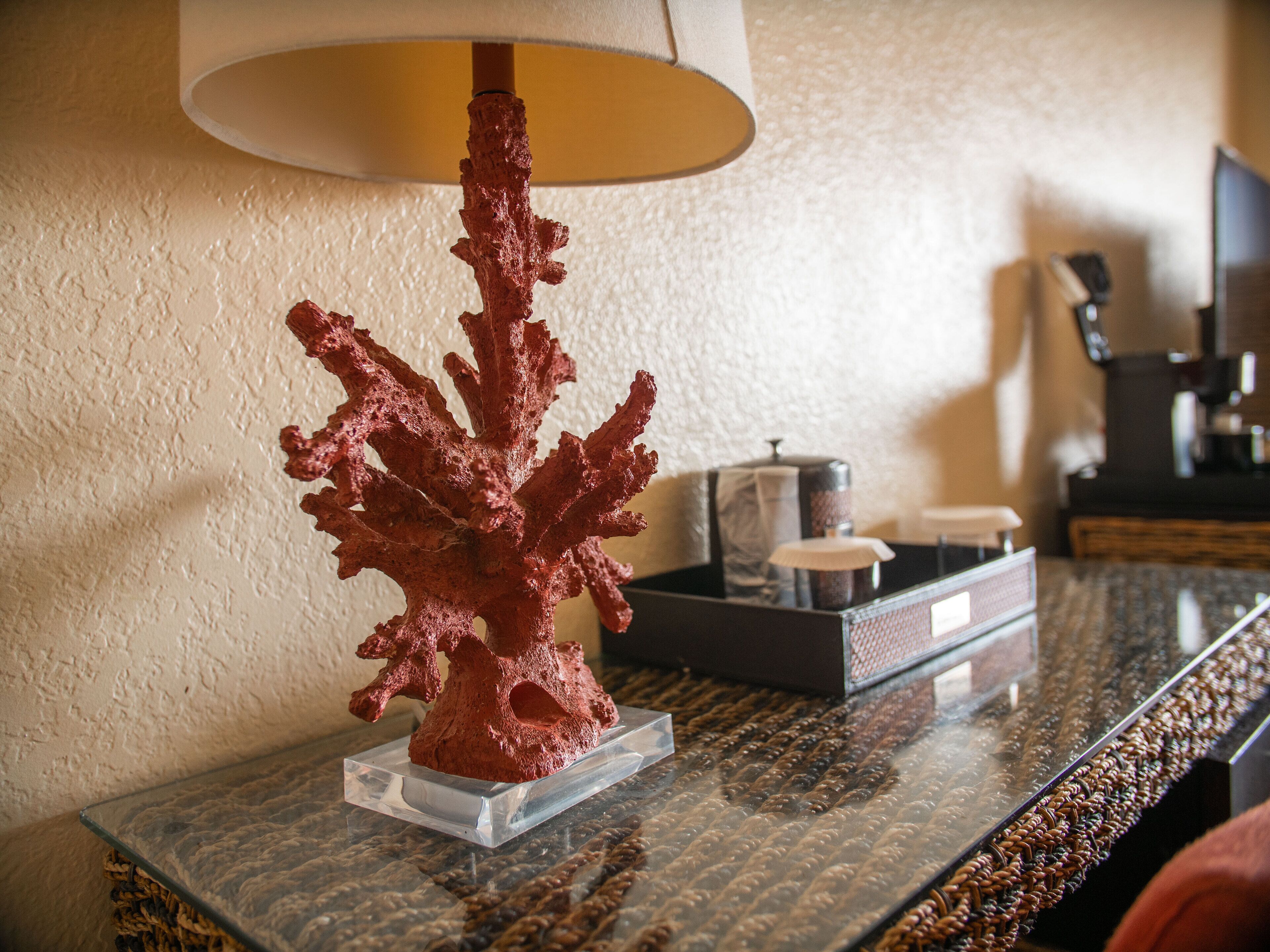 A coral sculpture on a clear glass stand on a marble table, with a lamp and a tray of items in the background.