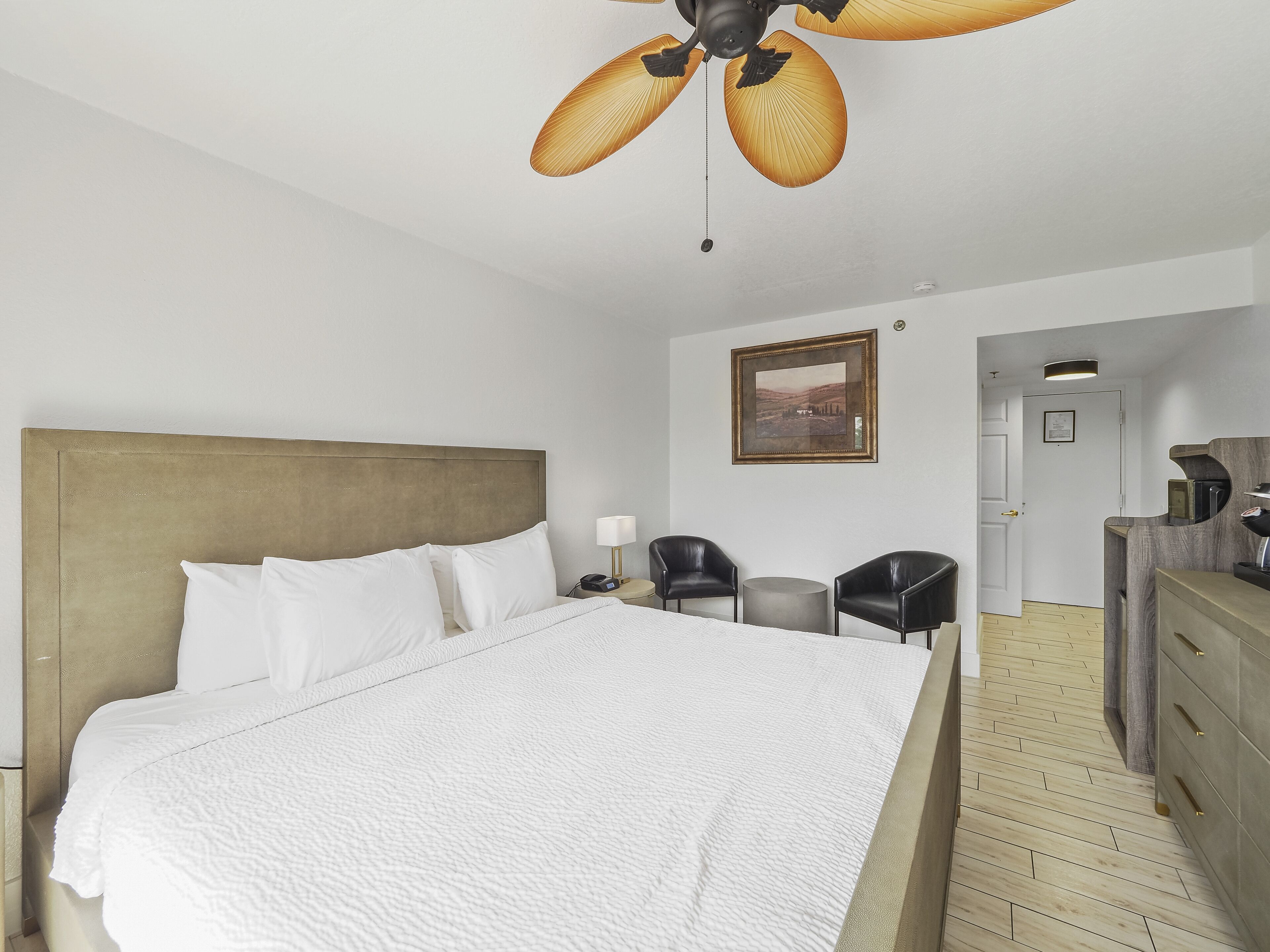 A hotel room with a large bed, a ceiling fan, a desk with a chair, and a door leading to another room.