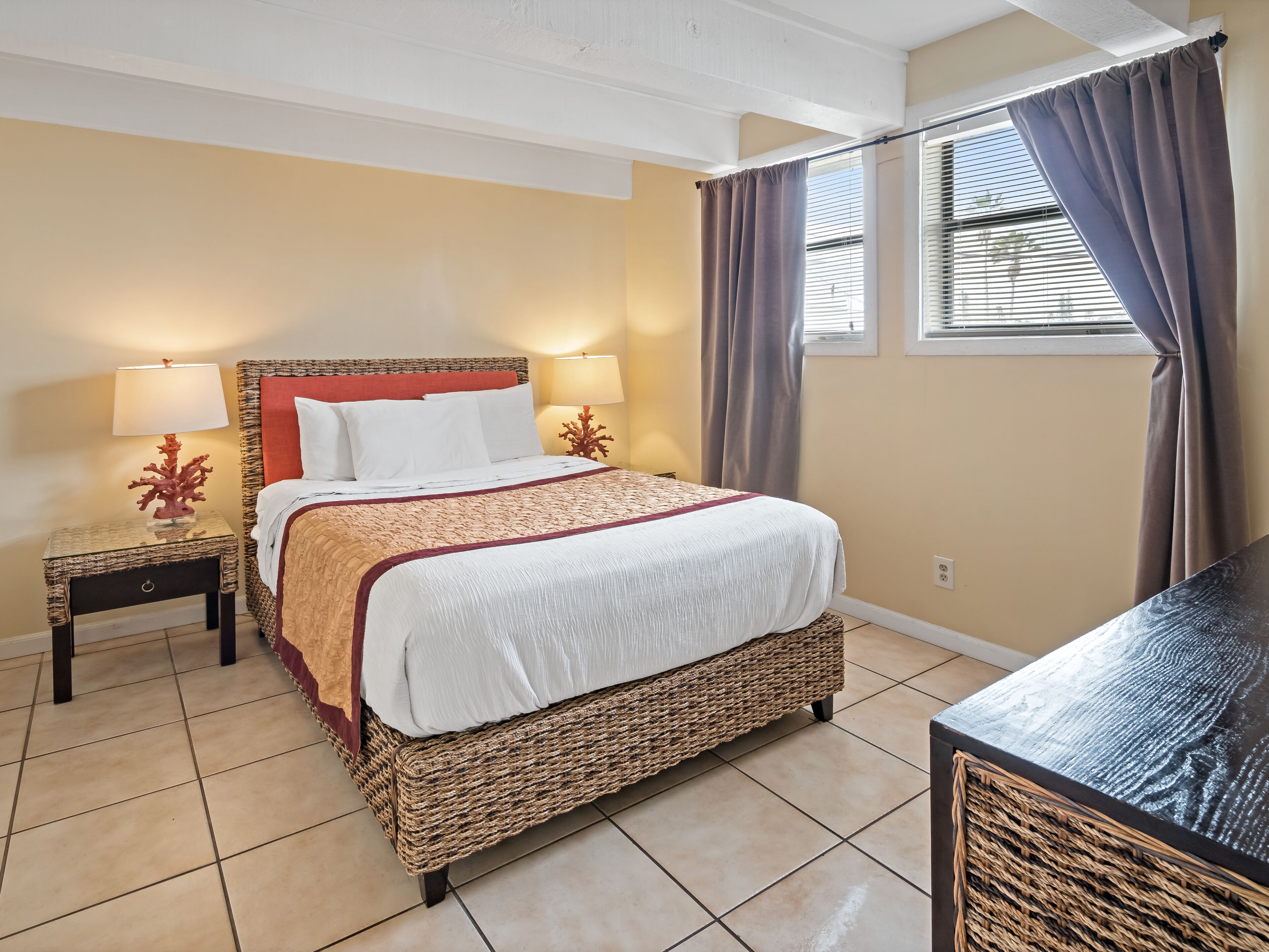 A hotel room with a large bed, two bedside tables with lamps, a wicker chair, and a window with curtains.