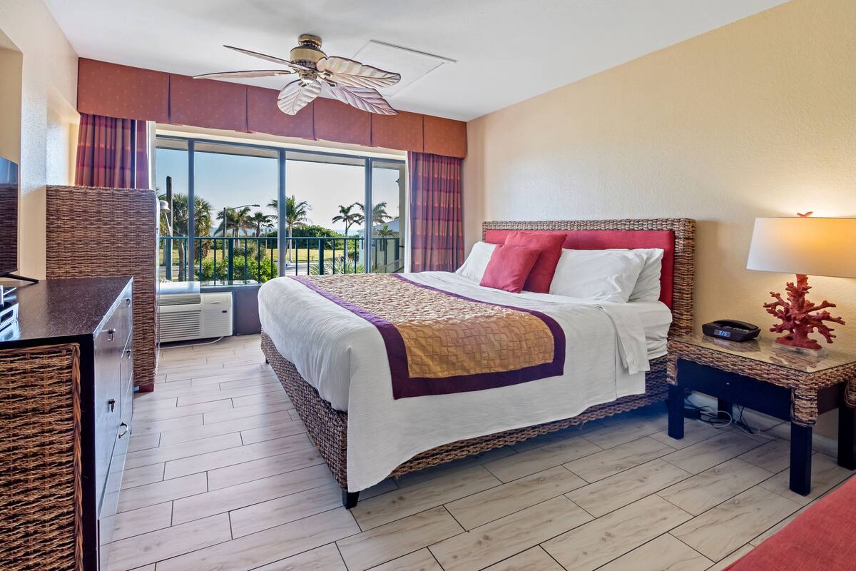 A hotel room with a large bed, a ceiling fan, a view of palm trees, and a balcony.