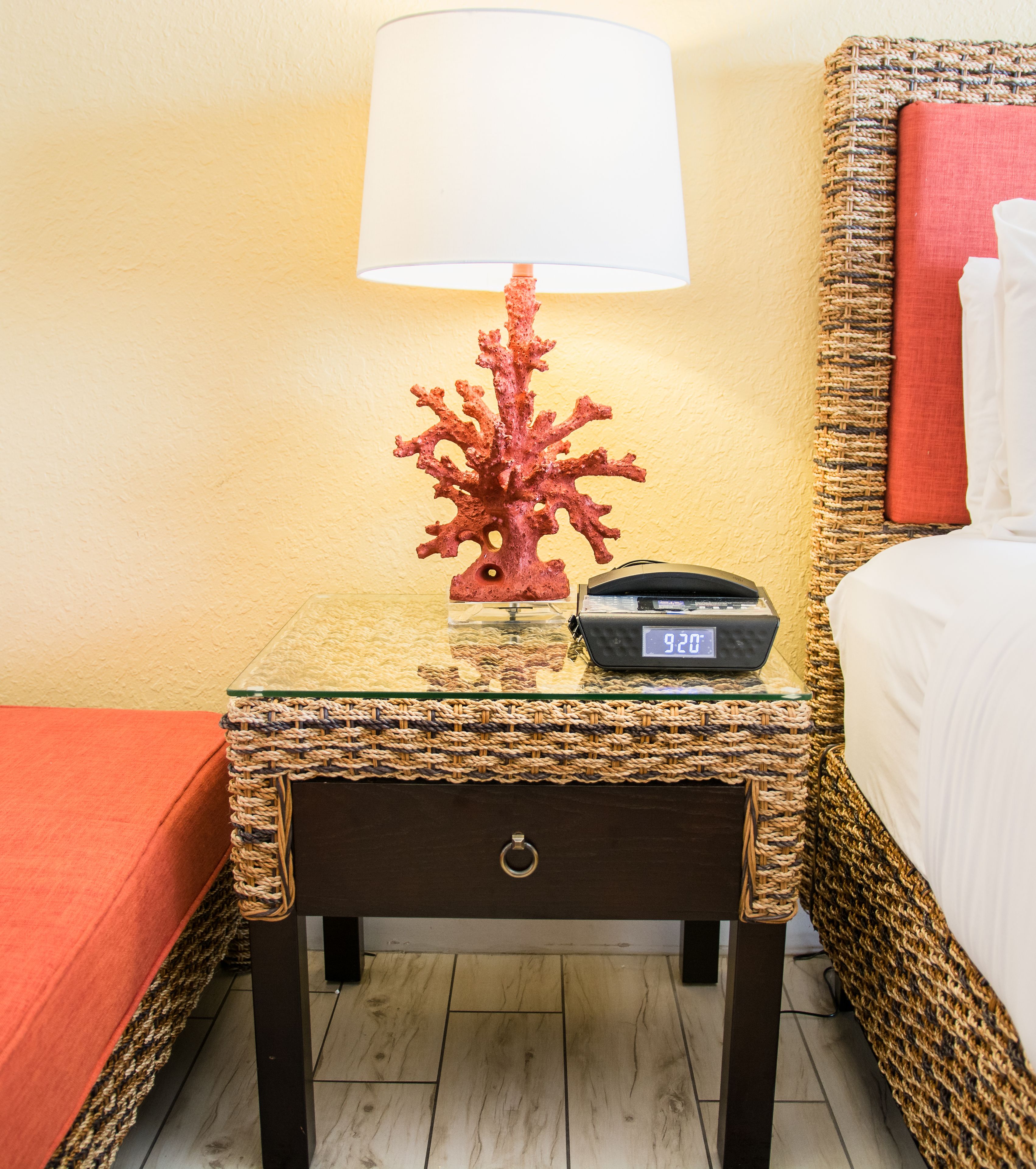 Une chambre d’hôtel avec une table de chevet en osier, un objet de décoration corail, une horloge numérique et un lit avec une tête de lit rouge.