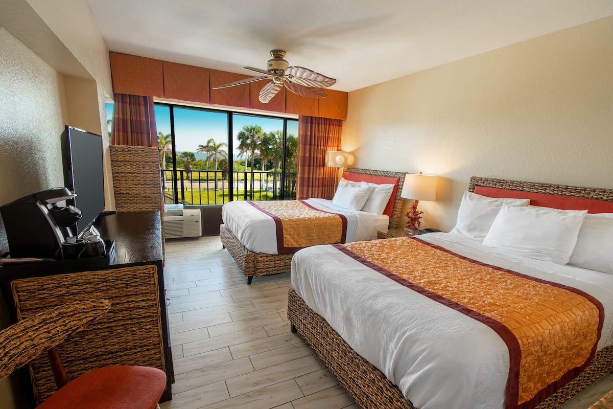 A hotel room with two beds, a desk with a coffee maker, a TV, and a balcony with a view of palm trees.