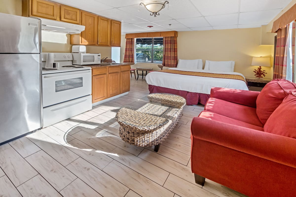 A compact hotel room with a kitchenette, a bed with a red headboard, a wicker chair, and a red sofa.