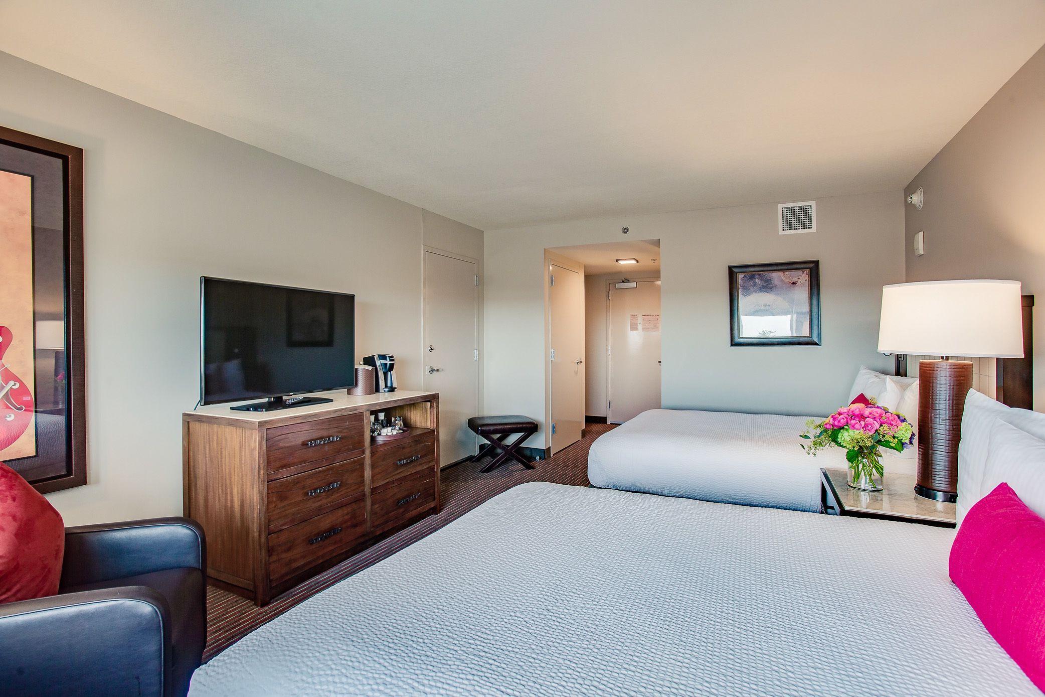 A hotel room with two beds, a TV, a dresser, and a desk with a lamp and flowers.