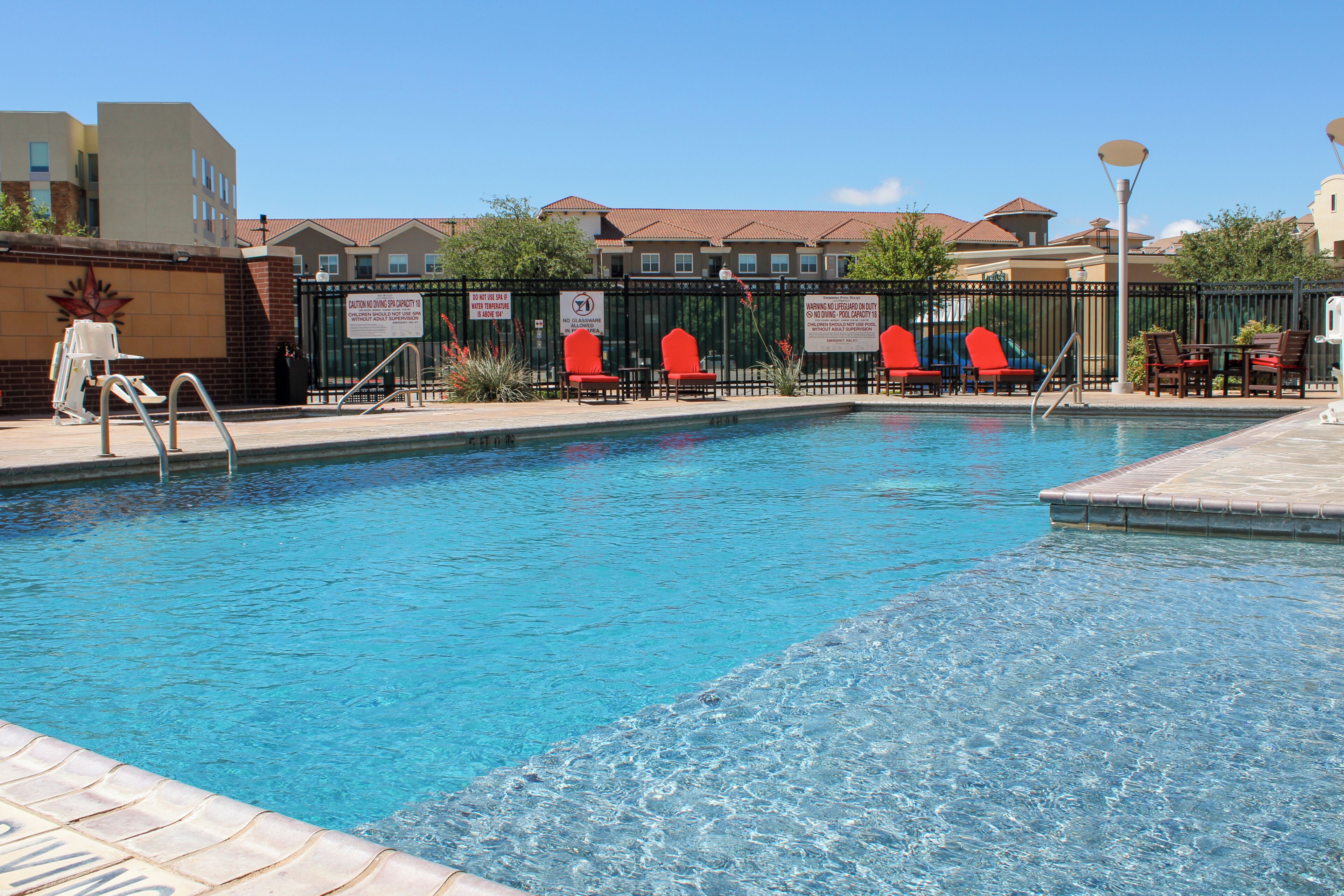 A swimming pool with clear water, surrounded by a fence, and red chairs along the edge.