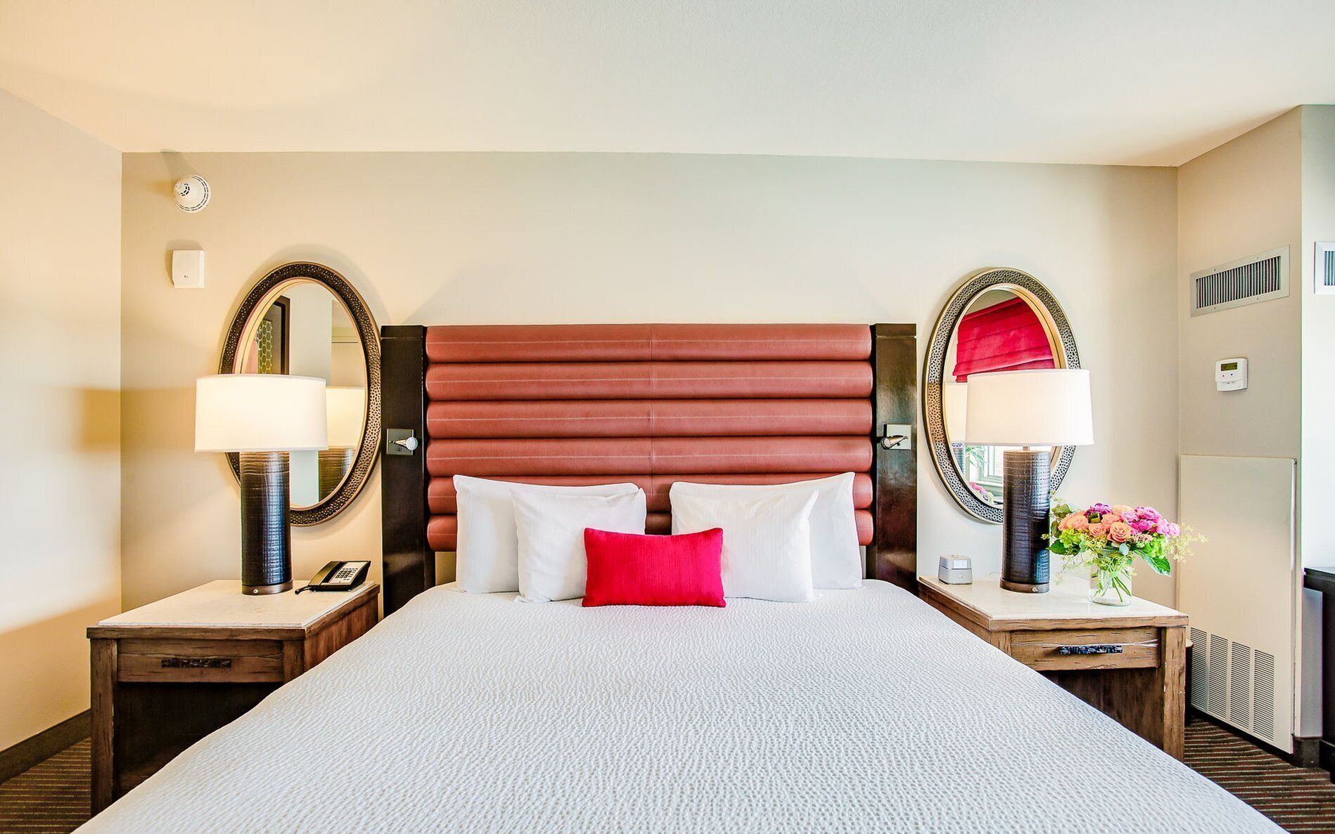 A hotel room with a large bed, two bedside tables, a headboard, two wall-mounted mirrors, a nightstand with a lamp, a thermostat, and a small plant.