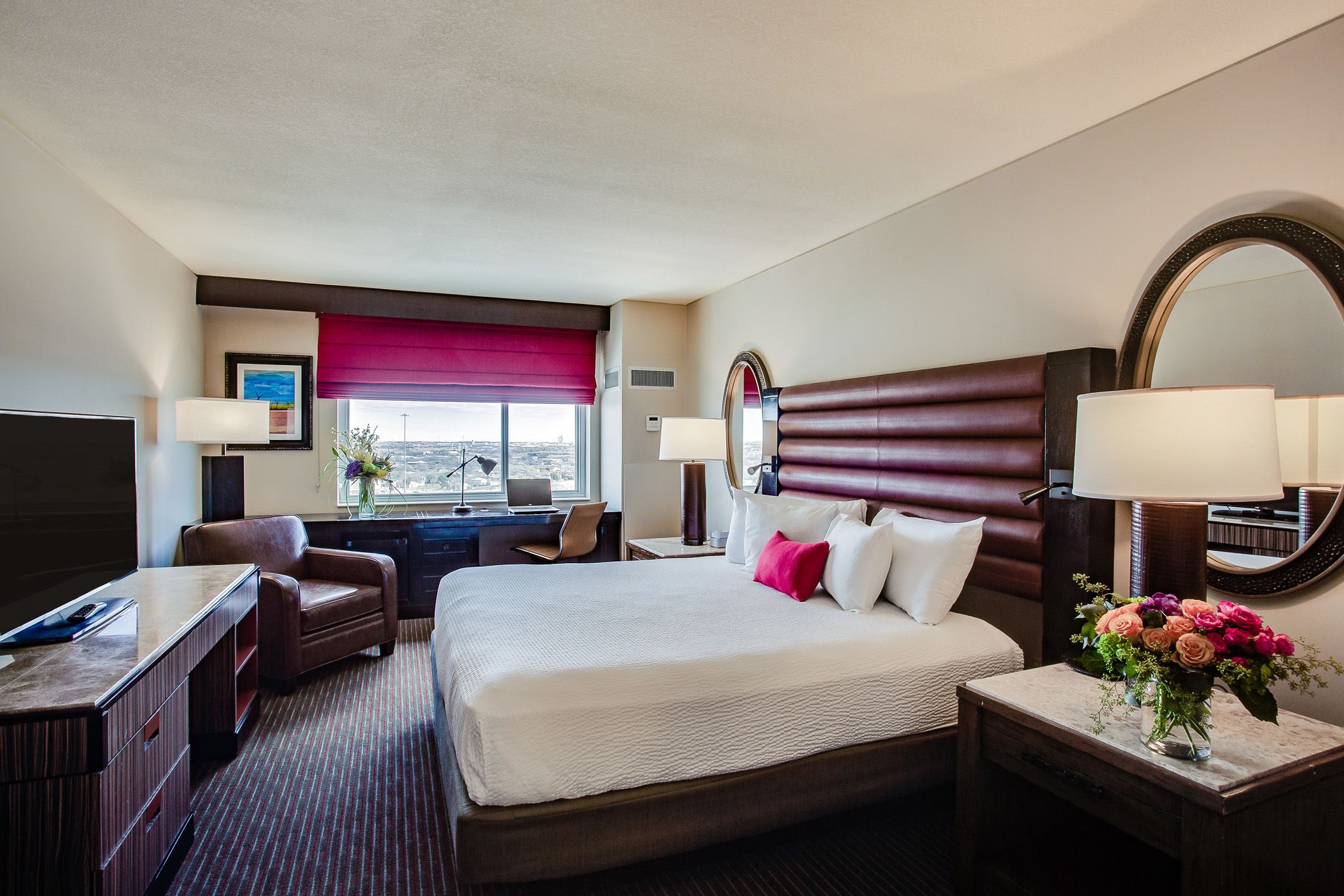 A hotel room with a large bed, a desk, a chair, a TV, and a window with a city view.