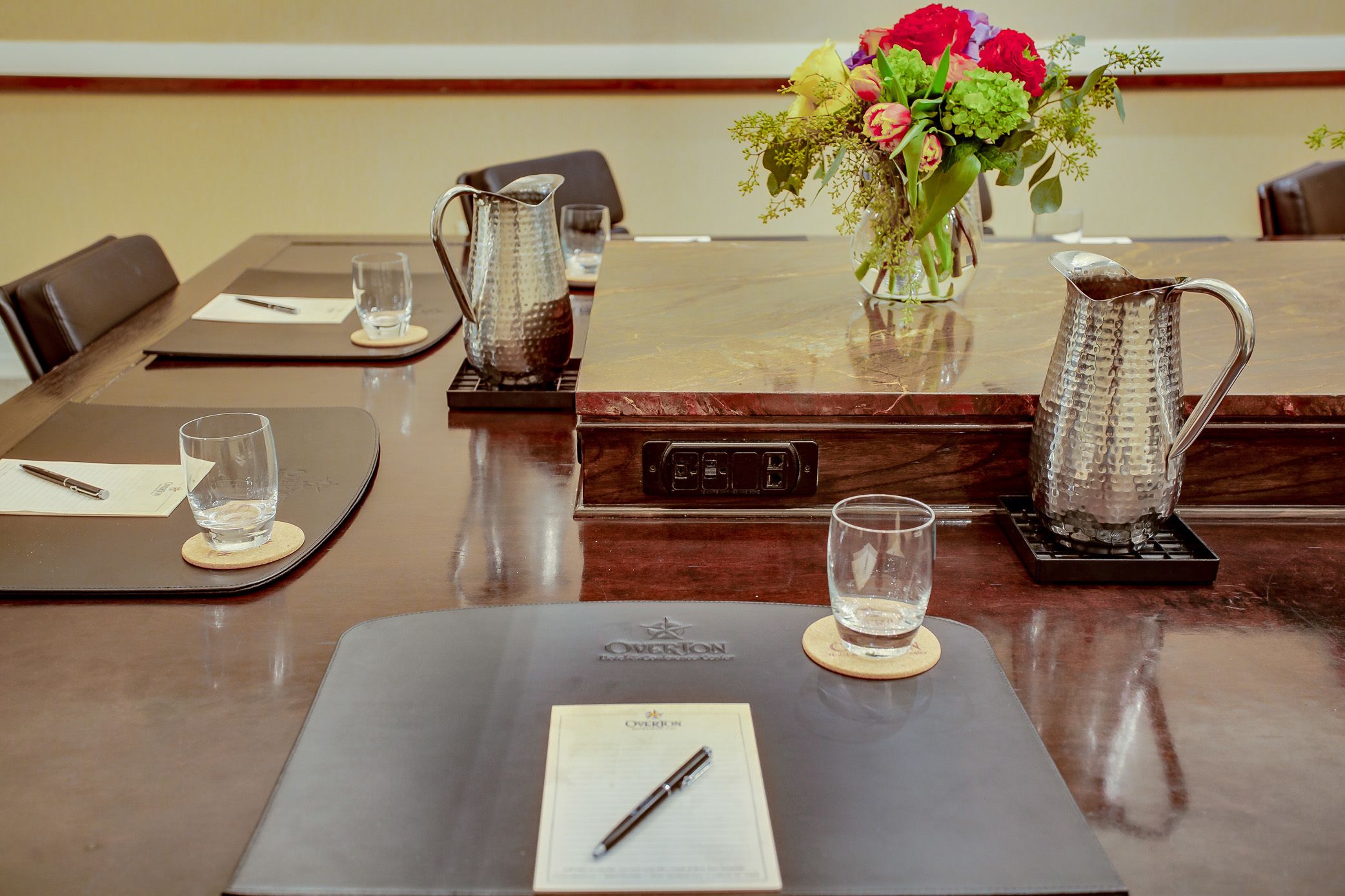 A conference table with a pitcher, glasses, a vase with flowers, and a nameplate.