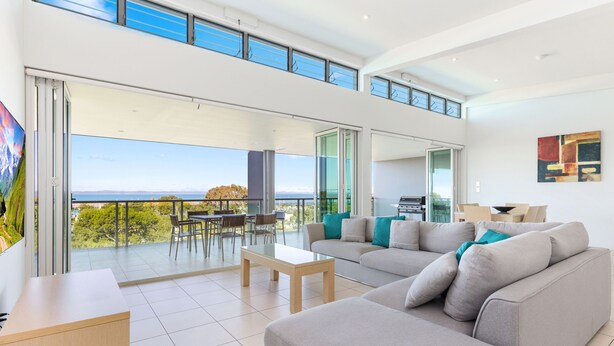 Uma sala moderna com um sofá cinzento grande, uma mesa de centro de madeira e um espaço de jantar com cadeiras. Há uma varanda com vista para o mar e uma área com churrasqueira.