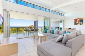 Una sala de estar moderna con un sofá grande de color gris, una mesa de centro de madera y un comedor con sillas. Hay un balcón con vistas al mar y una zona de barbacoa.