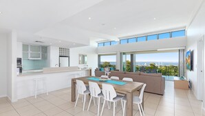 Cozinha moderna com armários brancos e uma ilha grande, sala de jantar com mesa e cadeiras de madeira, e sala de estar com sofá e grandes janelas com vista panorâmica.