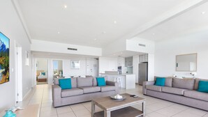 Uma sala de estar moderna com dois sofás cinzas, uma mesa de centro de madeira e uma cozinha ao fundo.