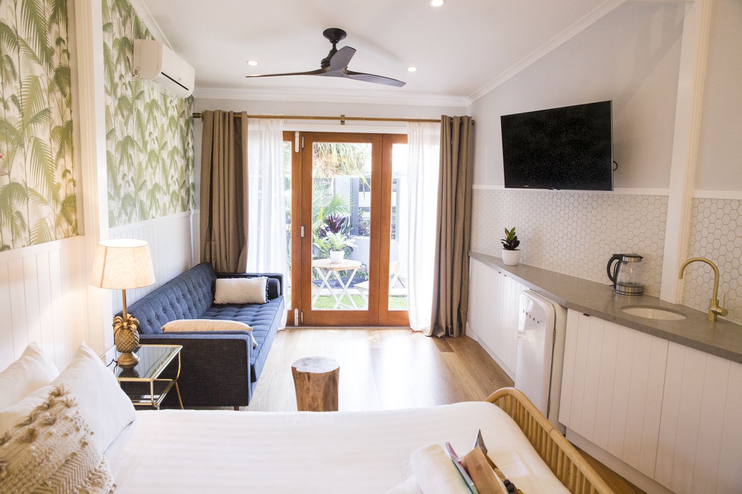 A hotel room with a bed, a sofa, a small table with a lamp, a kitchen area with a sink and a TV mounted on the wall.
