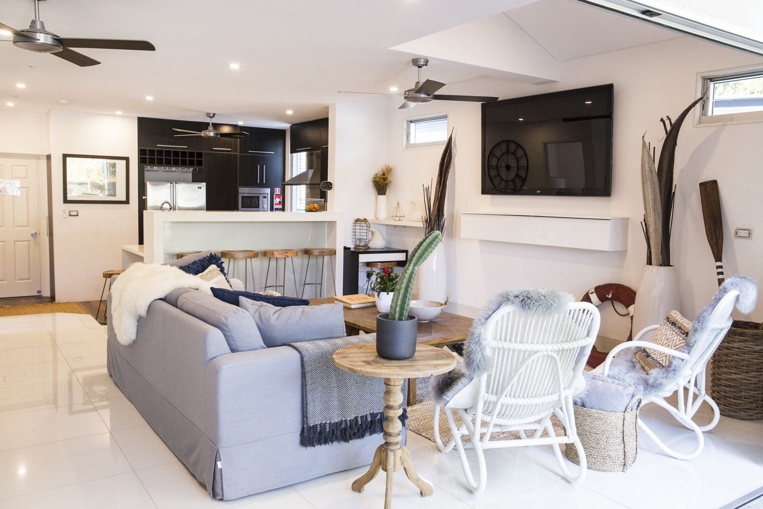 A modern living room with a grey sofa, white wicker chairs, a round wooden coffee table, and a kitchen in the background.