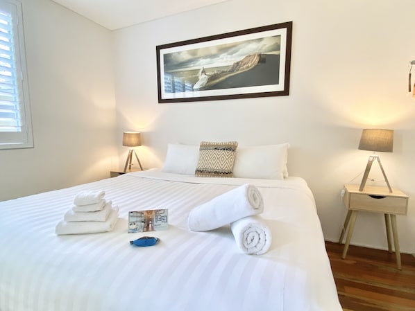 A neatly made bed with white linens, a rolled towel, and a blue item on the bed. There is a framed picture on the wall above the bed. - Bay Haven Lodge (Byron Bay)