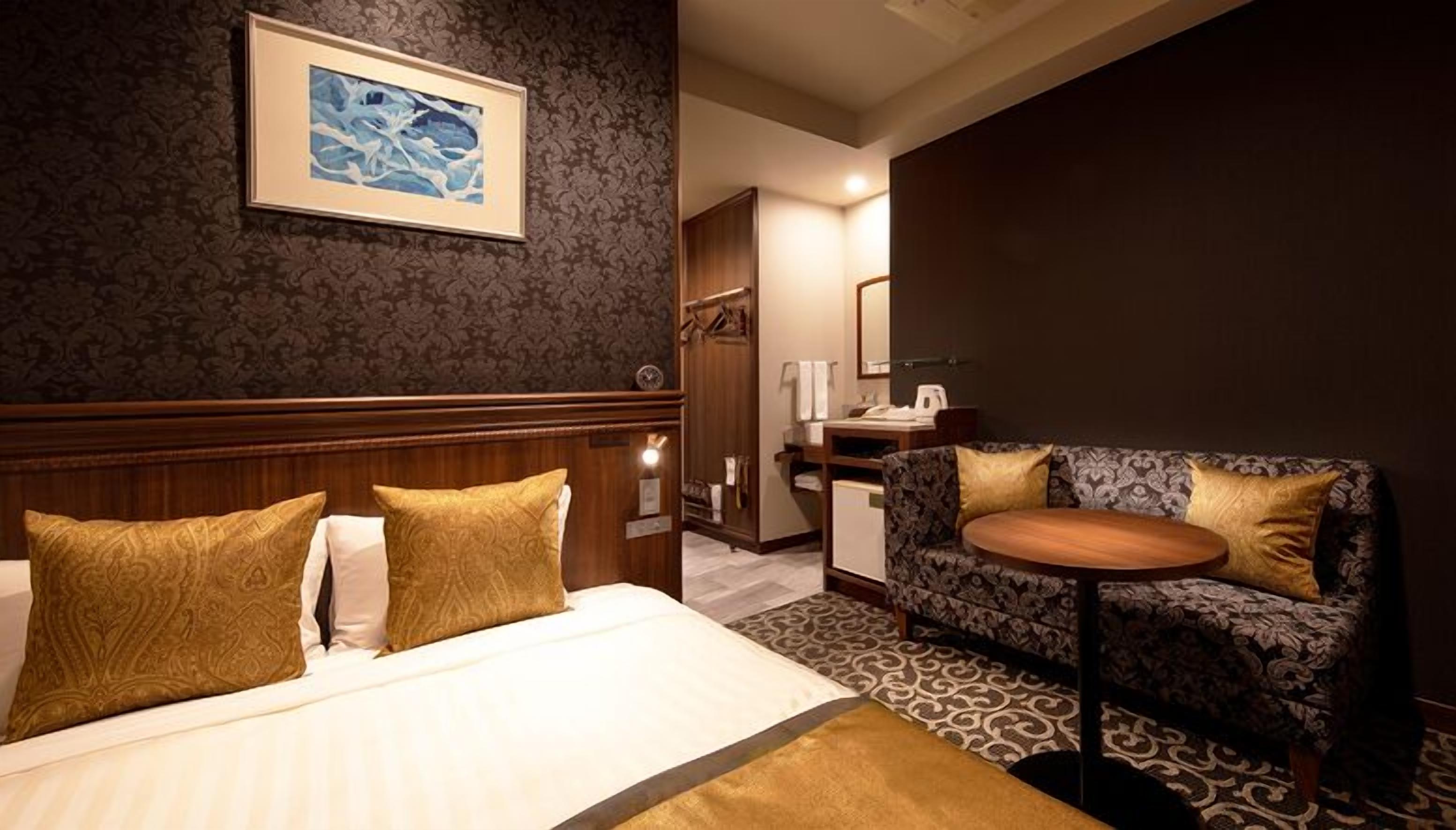 Habitación de hotel con cama, dos almohadas con diseño dorado, un cuadro enmarcado en la pared, un sofá con un cojín estampado y una pequeña mesa redonda.