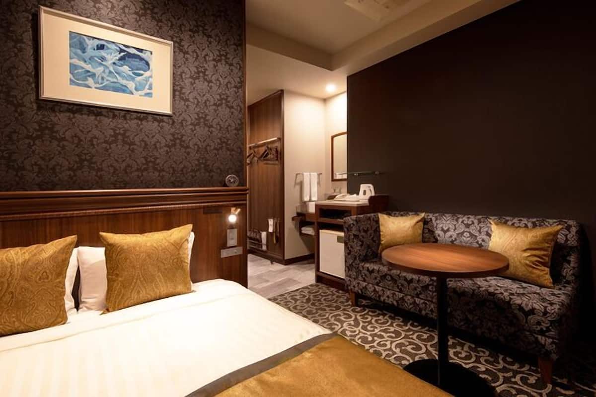 A hotel room with a bed, two gold-patterned pillows, a framed artwork on the wall, a sofa with a patterned cushion, and a small round table.