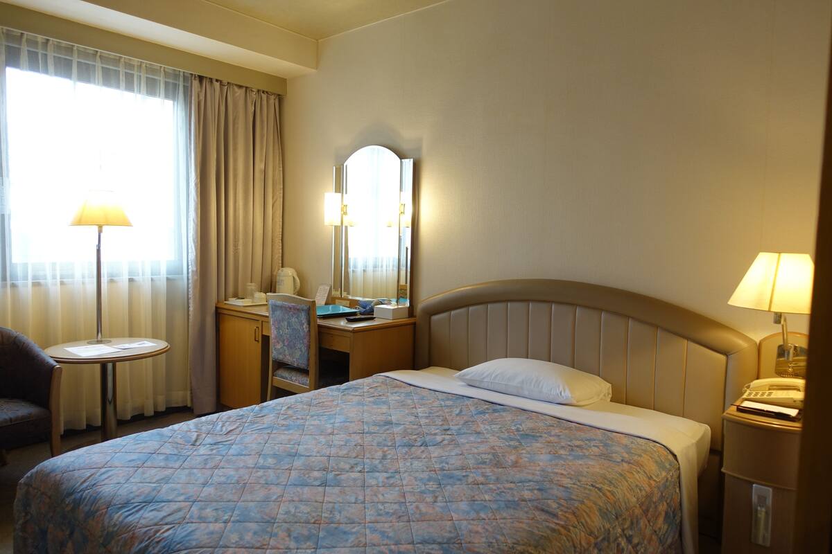 A hotel room with a bed, a desk, a chair, a lamp, and a mirror.