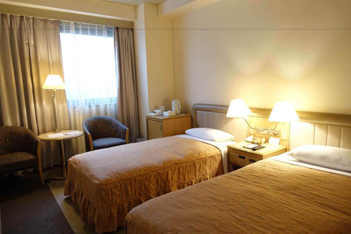 A hotel room with two beds, a chair, a table, and a nightstand with lamps.