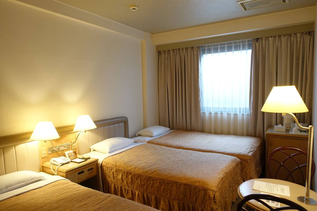 A hotel room with two beds, a desk with a lamp, a chair, and a window with curtains.
