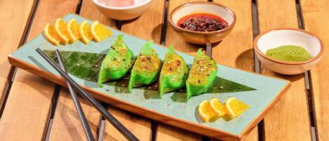 Un plato de dumplings verdes con rodajas de naranja y dos recipientes pequeños de salsa para mojar sobre una mesa de madera.