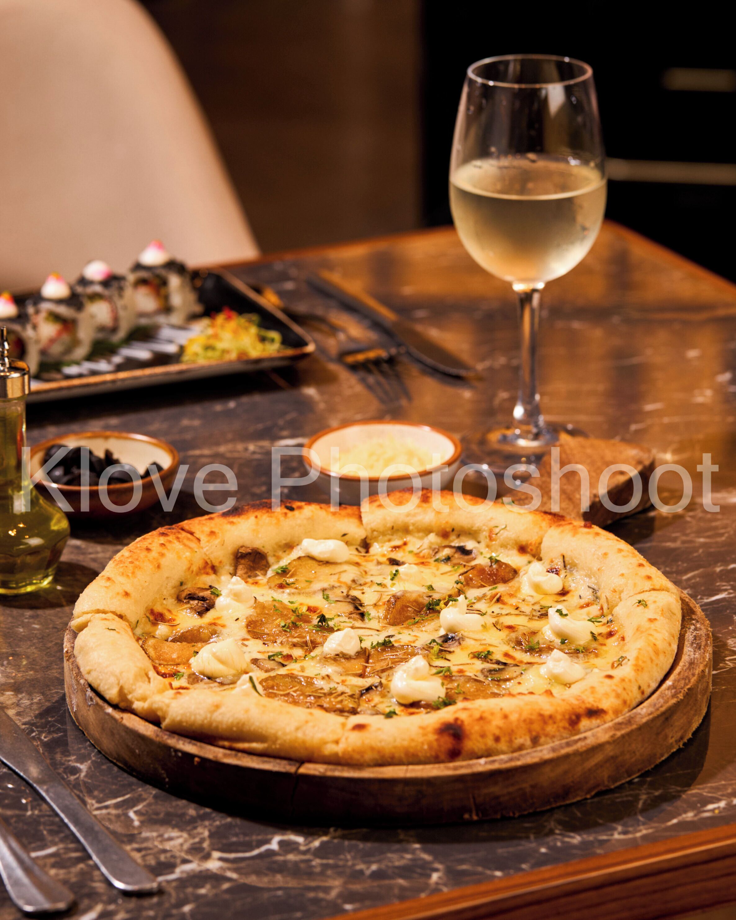 A pizza with various toppings on a wooden board, a glass of white wine, and a plate of sushi in the background.