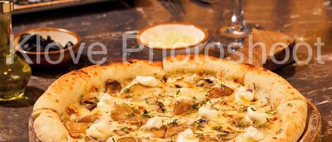 A pizza with various toppings on a wooden board, a glass of white wine, and a plate of sushi in the background.