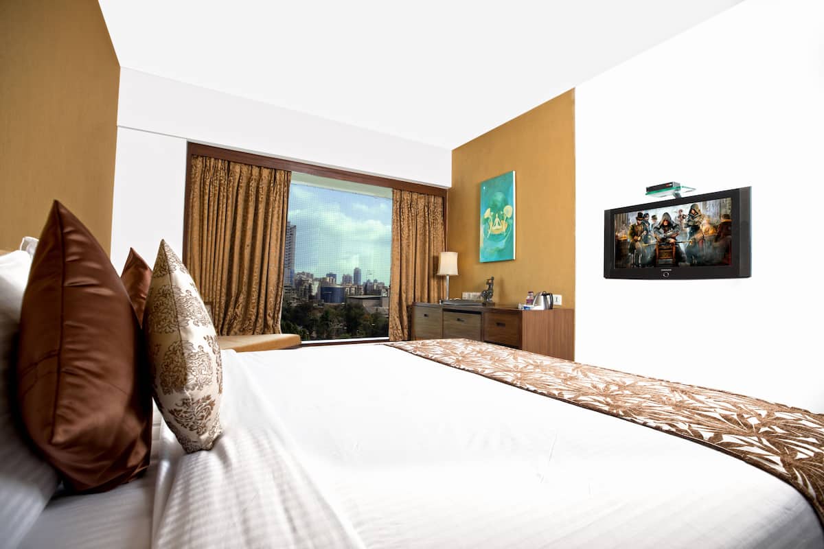 A hotel room with a large bed, a city view, a desk with a lamp, and a television on the wall.