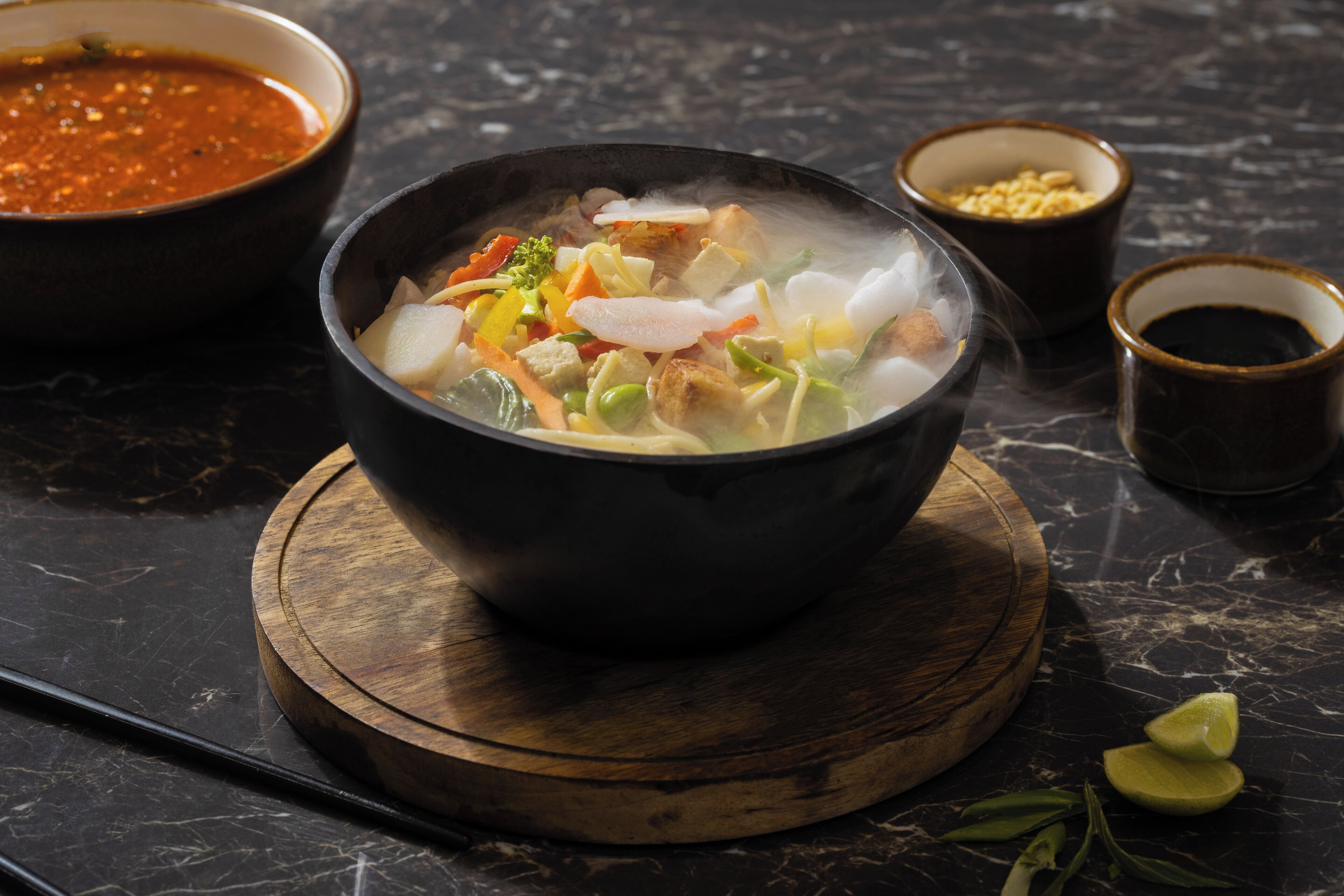 A bowl of soup with various vegetables and meat on a wooden board, accompanied by a bowl of sauce and two small cups on a dark marble surface.