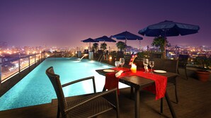 Area piscina sul tetto con tavolo apparecchiato per cenare, circondata da ombrelloni e sedie, con vista sulla città illuminata di notte.