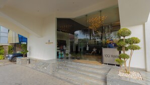 Ingresso di un hotel con reception, una scalinata che conduce alla hall e un'area parcheggio con un'auto blu e piante in vaso.