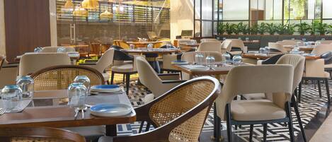 A restaurant with multiple tables and chairs, large windows, and indoor plants.