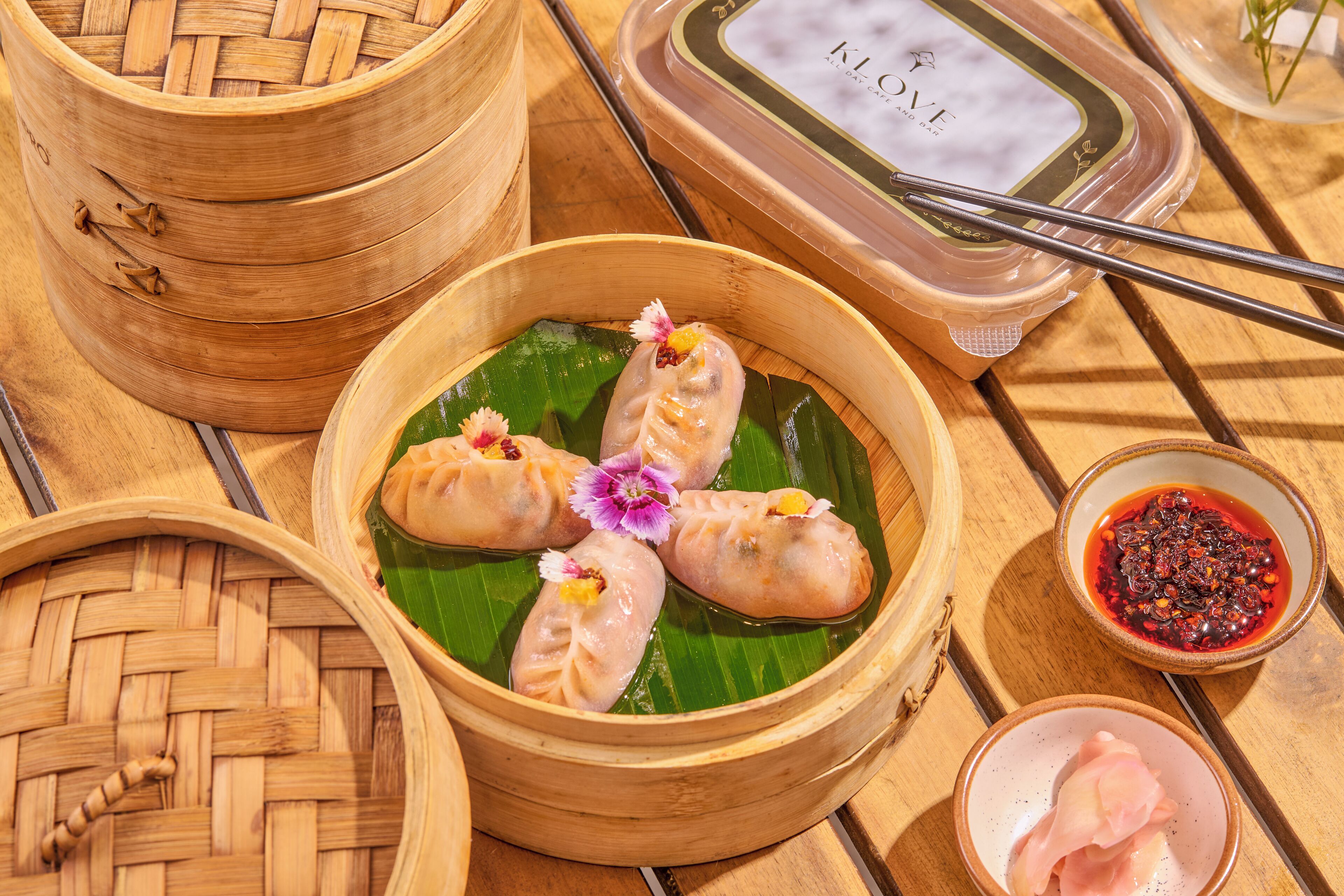 A bamboo steamer with dumplings on a banana leaf, a small bowl of dipping sauce, and a container of dipping sauce.