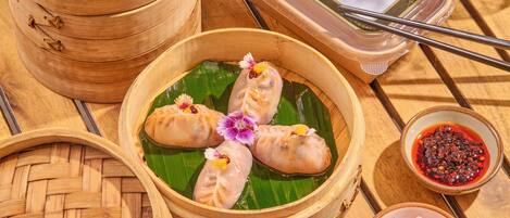 A bamboo steamer with dumplings on a banana leaf, a small bowl of dipping sauce, and a container of dipping sauce.