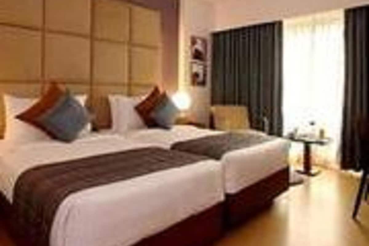 A hotel room with two beds, a large window with curtains, and a small table with a lamp.