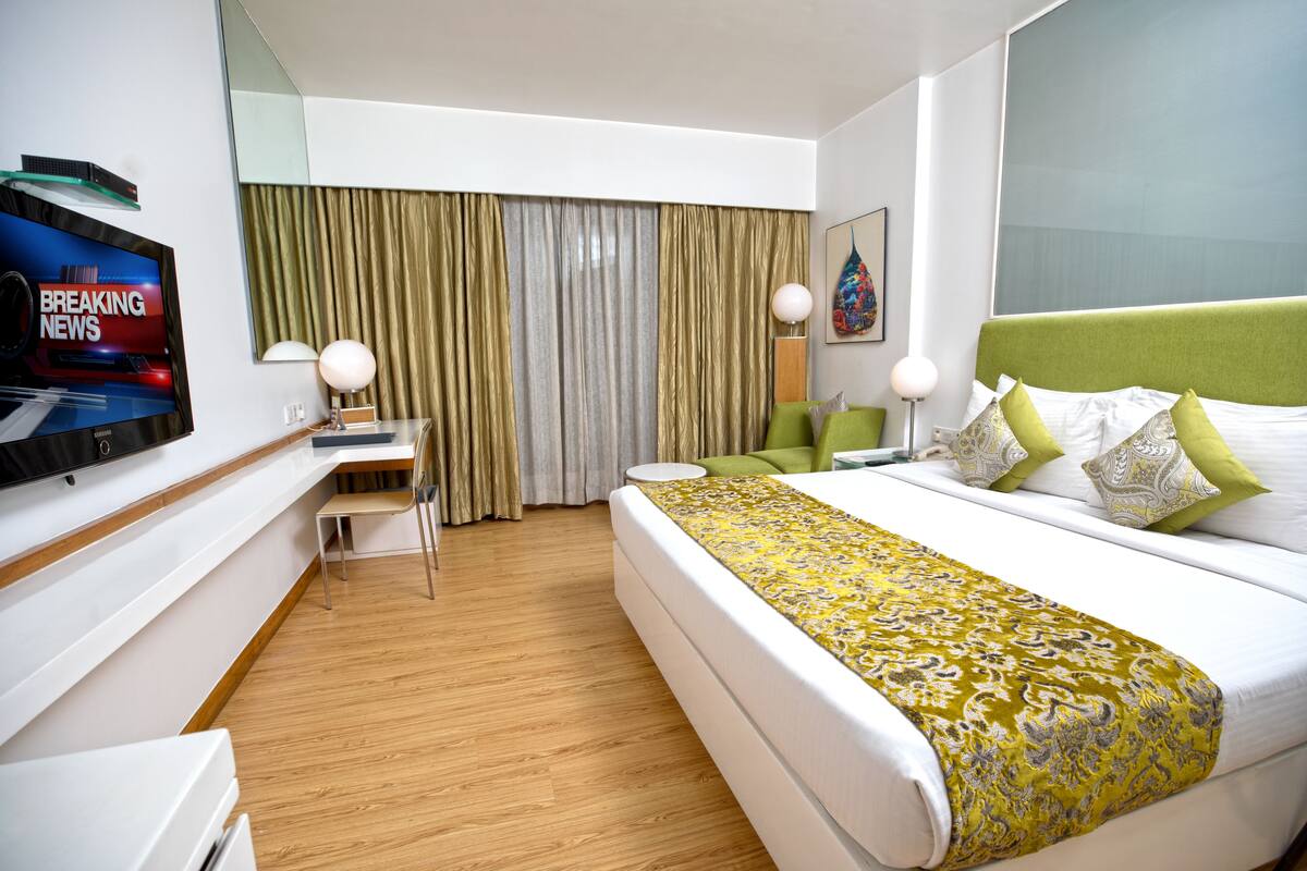 A modern hotel room with a large bed, a desk, a chair, a television, and a large window with curtains.