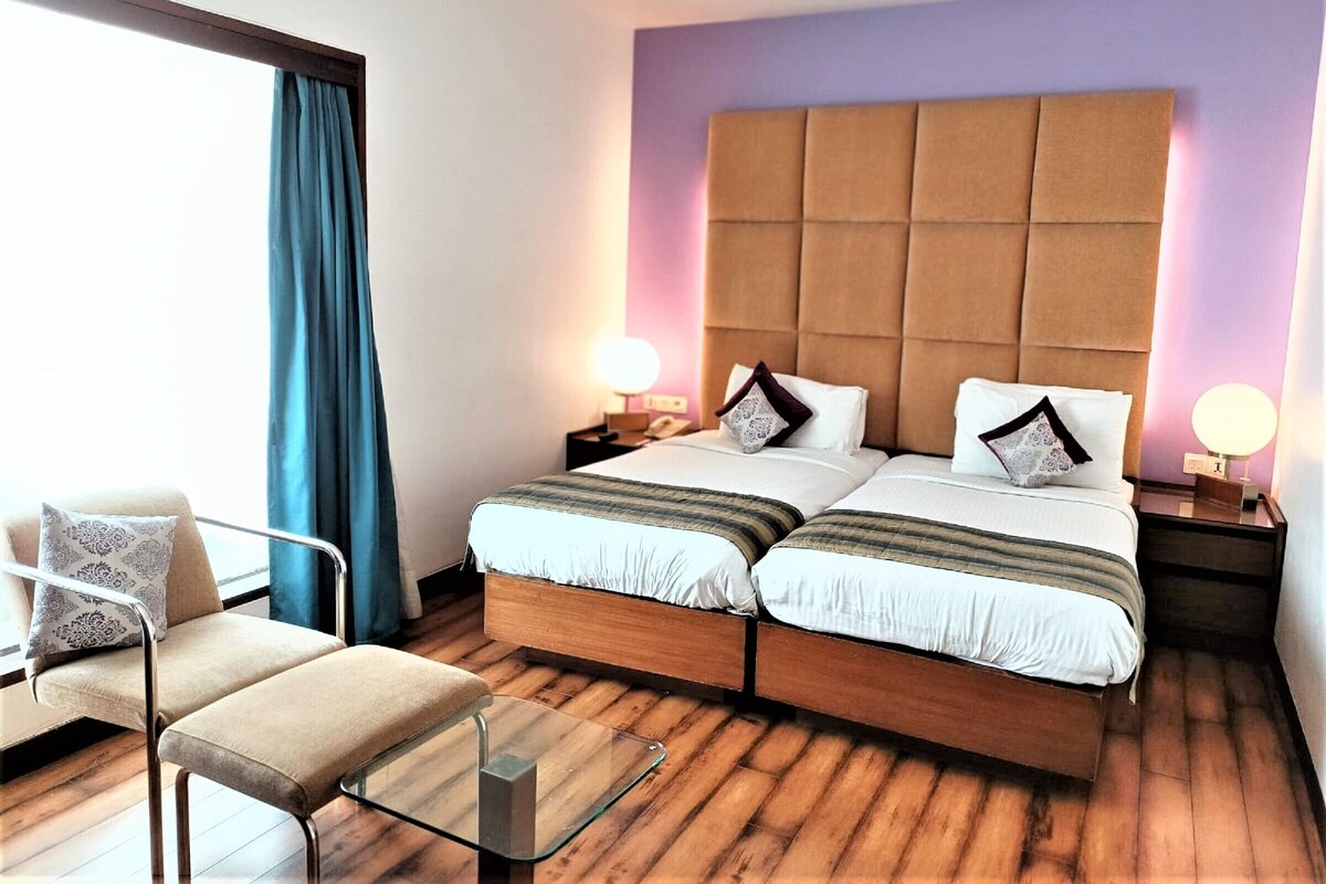 A double bed with a padded headboard, two bedside tables with lamps, a chair, and a window with curtains.