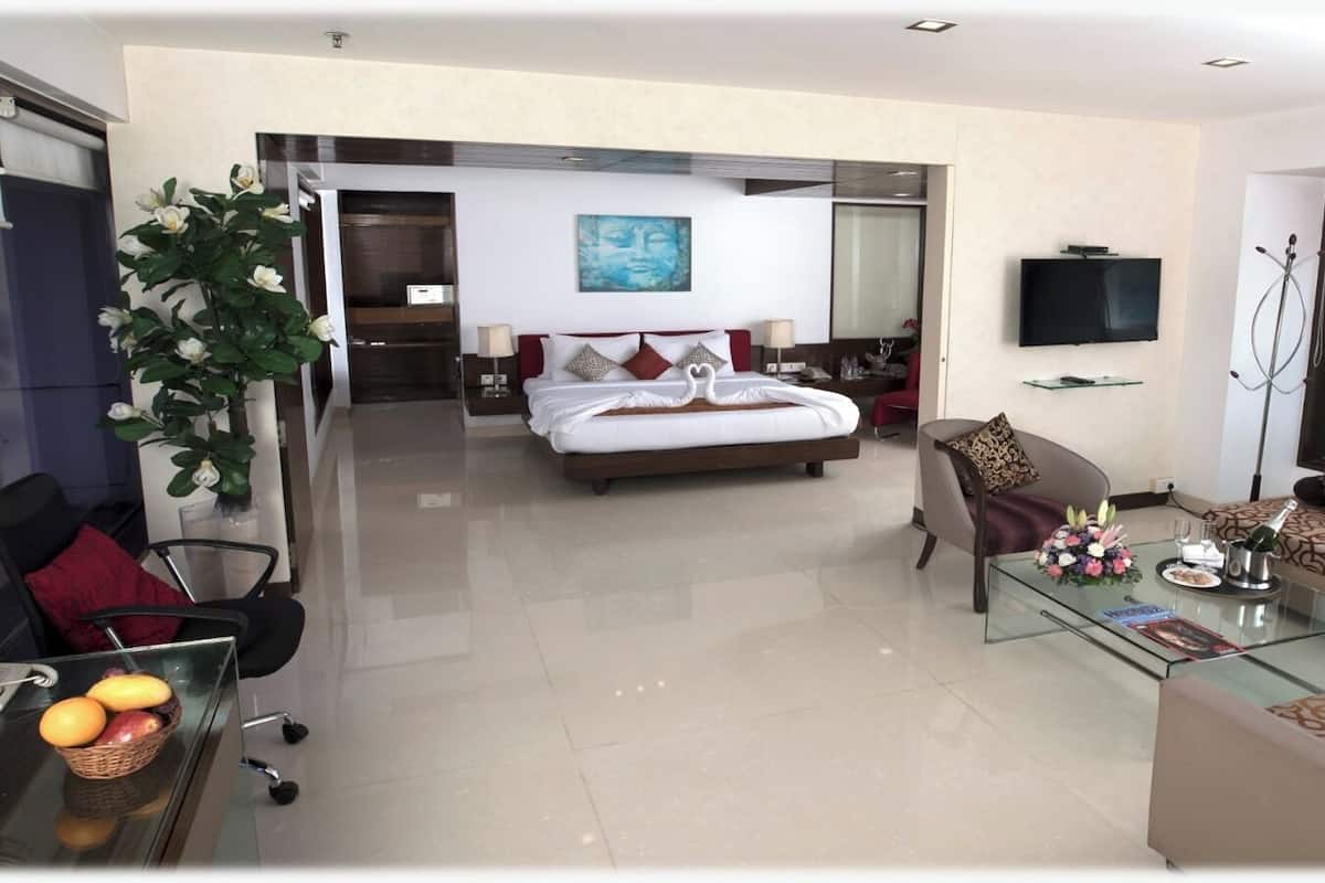 A modern hotel room with a bed, a TV, a glass coffee table, and a dining area with a basket of fruit.