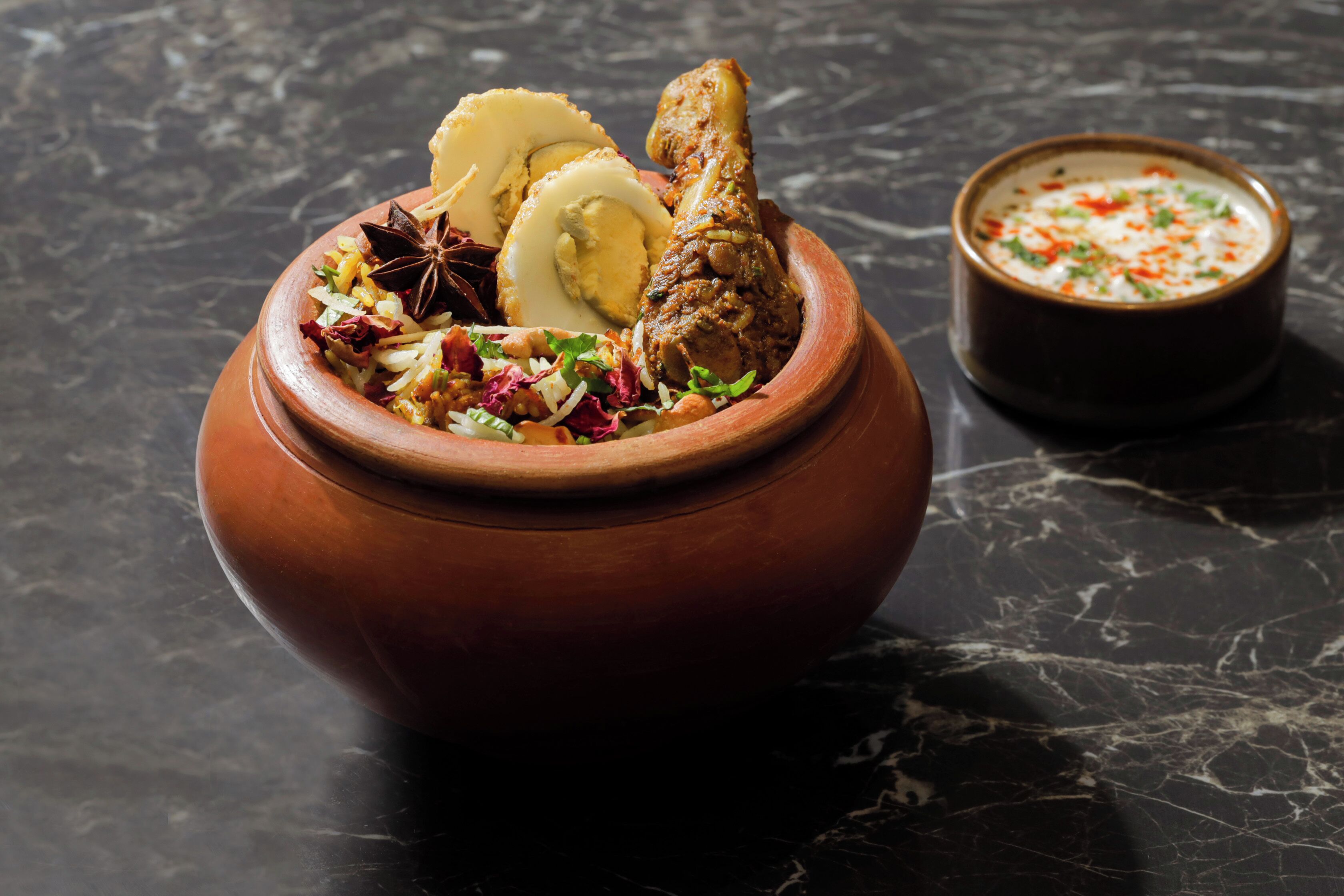 A clay pot with food and a small bowl of sauce on a dark marble surface.