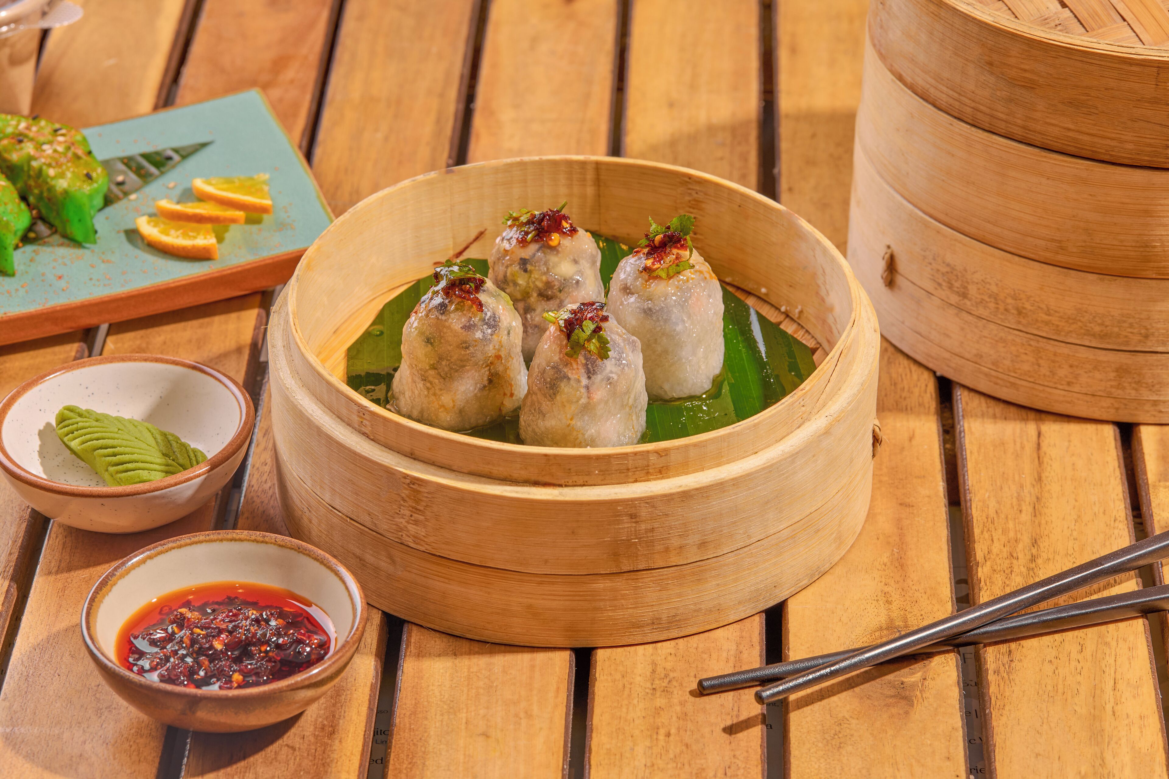 Una mesa de madera con una vaporera de bambú llena de dumplings, un tazón de salsa para mojar y un plato con fruta cortada.