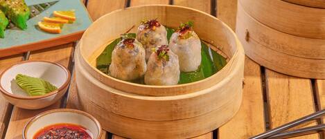 Una mesa de madera con una vaporera de bambú llena de dumplings, un tazón de salsa para mojar y un plato con fruta cortada.