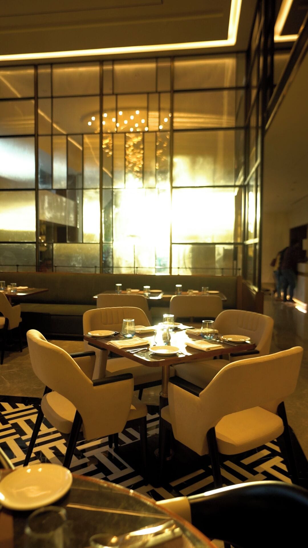 A modern restaurant interior with a central dining table surrounded by chairs, a patterned floor, and large windows allowing natural light.