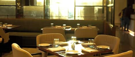 A modern restaurant interior with a central dining table surrounded by chairs, a patterned floor, and large windows allowing natural light.
